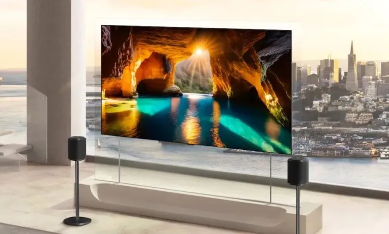 Transparent TV displays cave scene with city view behind, speakers on stands.
