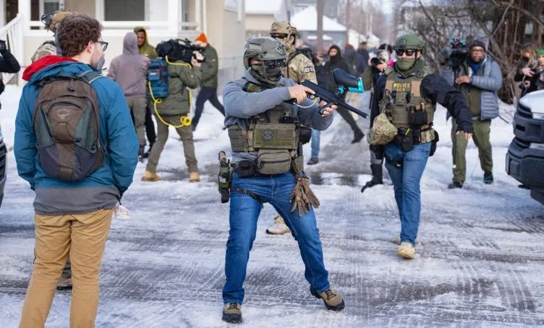 ICE officers with paintball guns surrounded by media on snowy street.