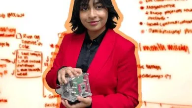 Young woman in red blazer holding a cubesat with whiteboard diagrams in background.