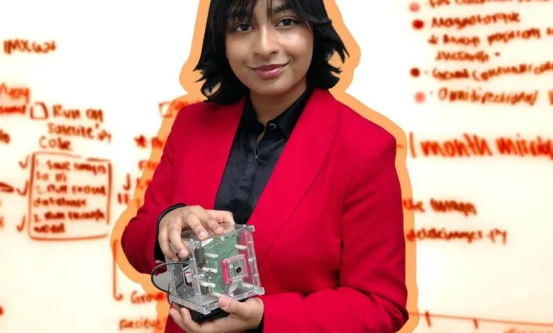 Young woman in red blazer holding a cubesat with whiteboard diagrams in background.