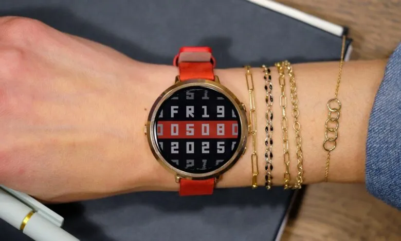 Wrist with gold smartwatch, red band, displaying date and gold bracelets.