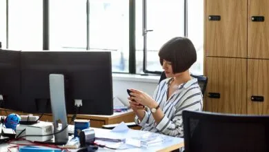 Woman with bob haircut using phone at messy office desk.