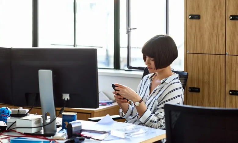 Woman with bob haircut using phone at messy office desk.