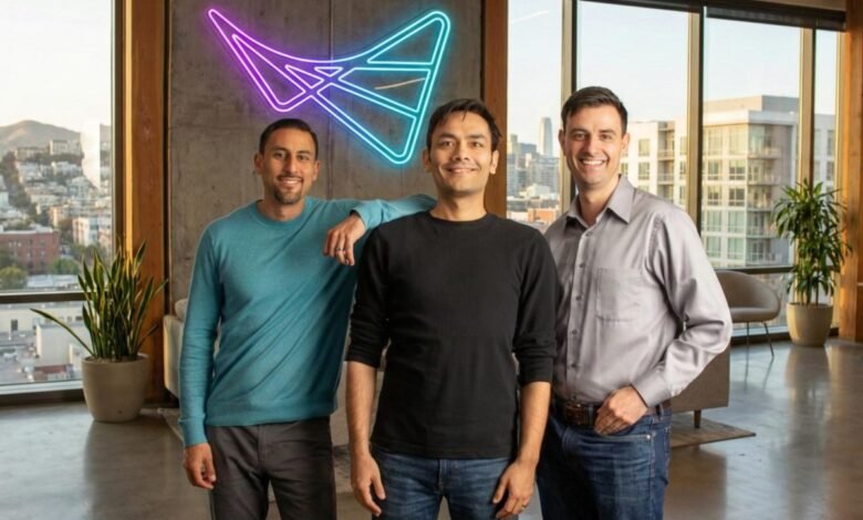 Three men stand in front of a window with a neon logo on the wall.
