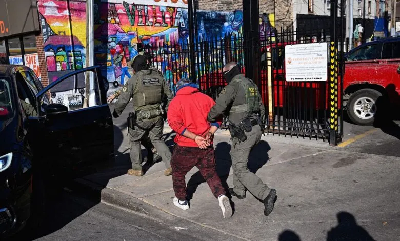 Man in handcuffs escorted by officers near a black car and colorful mural.
