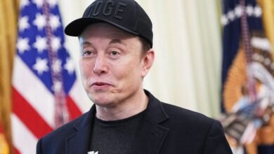 Elon Musk wearing a DOGE hat stands in front of US flags.