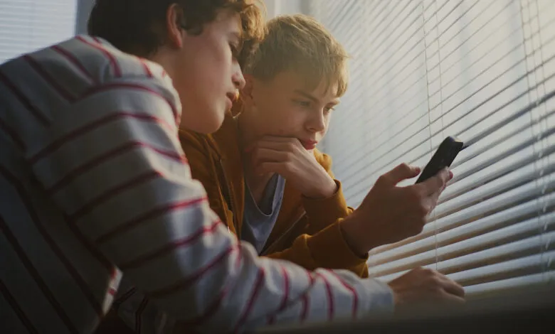 Two teenage boys looking at a phone by a window with blinds.