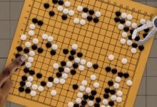 Go board with black and white stones, played by human and robotic hands.
