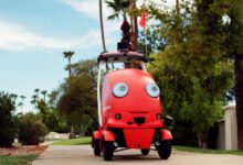 Red DoorDash delivery robot on sidewalk with palm trees and blue sky.