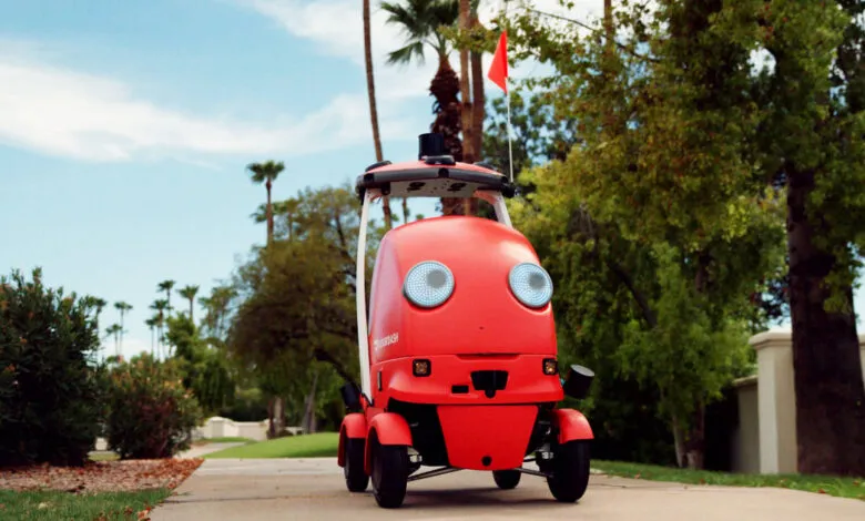 Red DoorDash delivery robot on sidewalk with palm trees and blue sky.