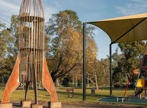 Metal rocket sculpture in a park with trees and a playground structure.