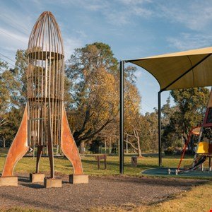 Metal rocket sculpture in a park with trees and a playground structure.