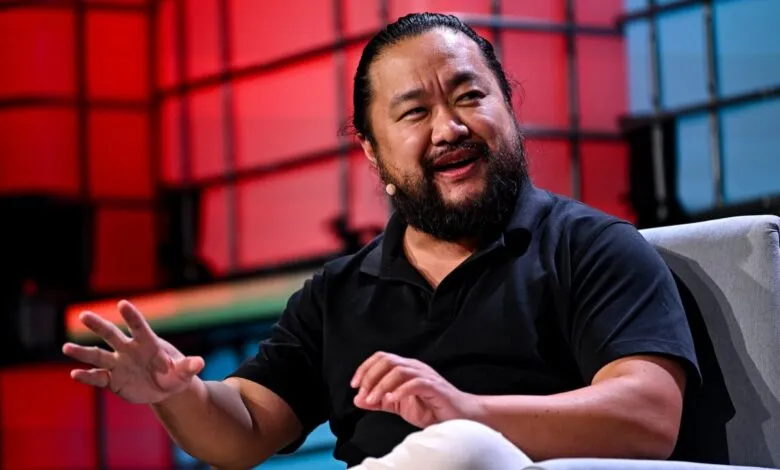Man with beard gestures while seated in chair on stage with red and blue background.