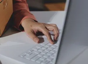 Hands typing on a white laptop next to a cardboard box, suggesting online business.