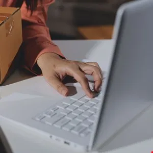 Hands typing on a white laptop next to a cardboard box, suggesting online business.