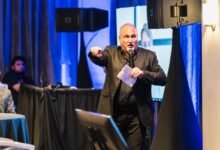 Man in black suit speaks into microphone on stage with blue lighting.