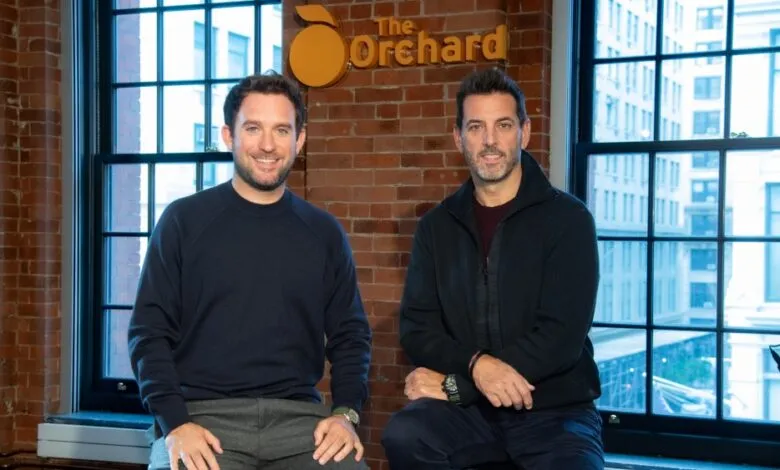 Two men pose in front of The Orchard logo, smiling at the camera.