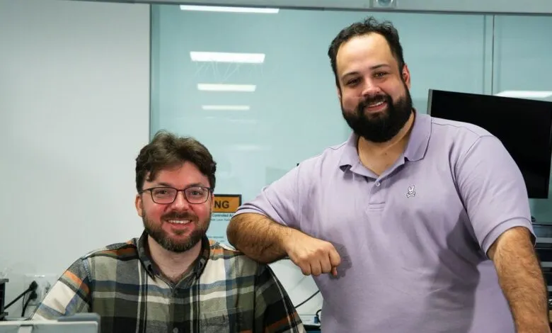 Two smiling men pose in an office setting, one leaning on the other.