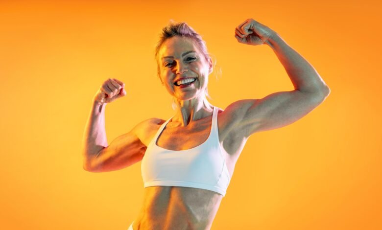 Smiling woman flexing biceps in white sports bra against orange background.