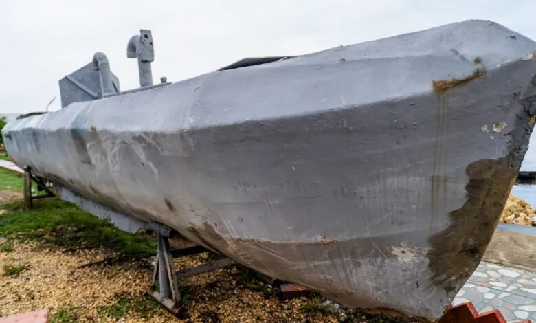 Gray submarine on land, supported by metal stands, near water.