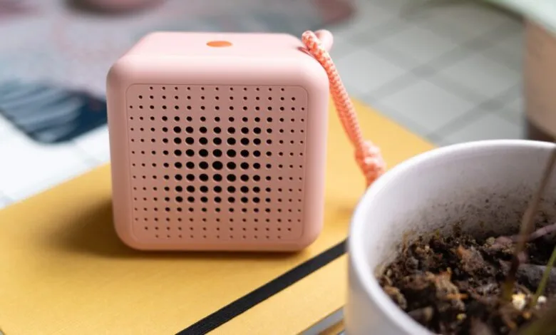 Pink cube speaker with holes on a yellow notebook, plant nearby.