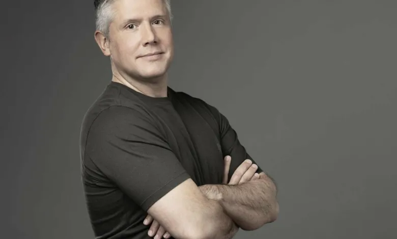 Headshot of actor Darren Mowry with arms crossed, smiling slightly against a gray background.