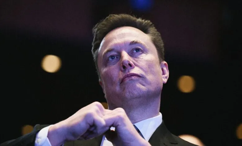 Elon Musk looking upwards with hands clasped, wearing a suit.