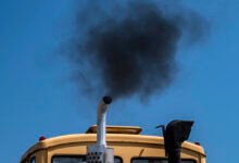 Black smoke billows from the exhaust pipe of a yellow truck against a blue sky.