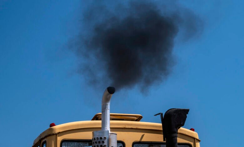 Black smoke billows from the exhaust pipe of a yellow truck against a blue sky.