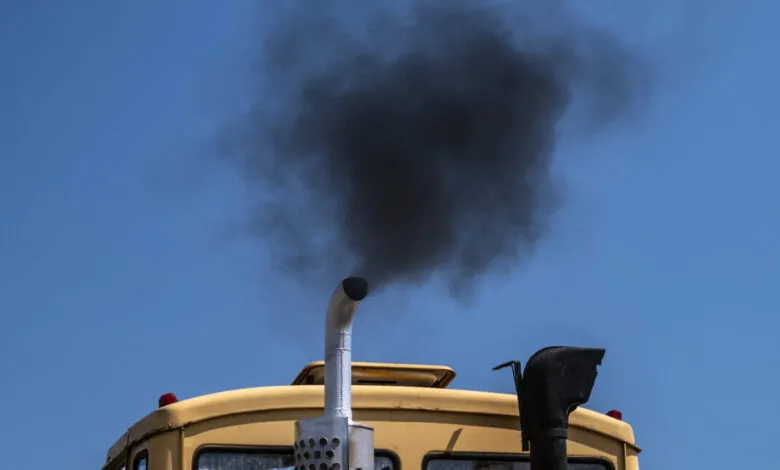 Black smoke billows from the exhaust pipe of a yellow truck against a blue sky.