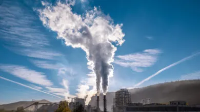 Industrial plant emits smoke from tall smokestacks into a blue sky with wispy clouds.