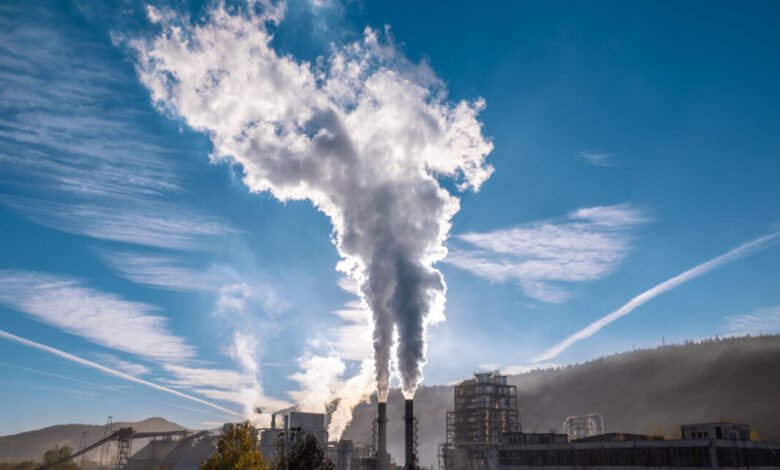 Industrial plant emits smoke from tall smokestacks into a blue sky with wispy clouds.