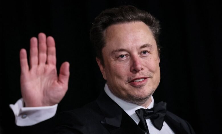 Elon Musk waves with a smile, wearing a tuxedo.