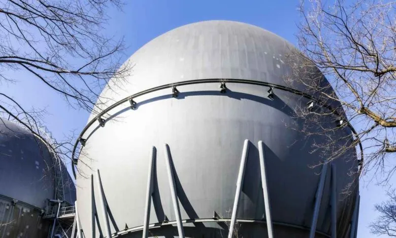 Two large, spherical gray gas holders against a clear blue sky, framed by bare tree branches.