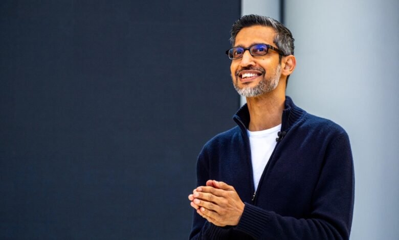 Sundar Pichai smiles warmly with hands clasped in front of a dark background.