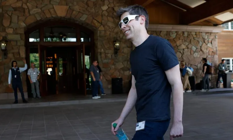 Sam Altman smiles wearing white glasses and holding a water bottle.