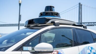 Waymo self-driving car with sensor suite on roof, San Francisco bridge in background.