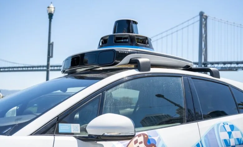 Waymo self-driving car with sensor suite on roof, San Francisco bridge in background.