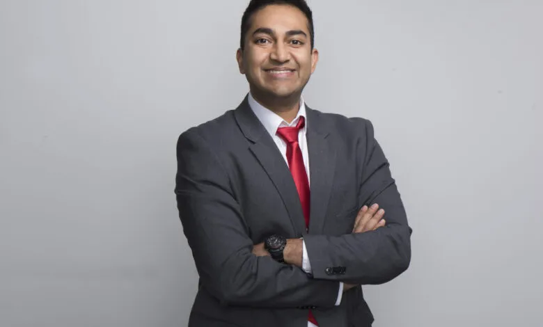 Dr. Vinay Prasad smiles confidently with arms crossed in a gray suit and red tie.