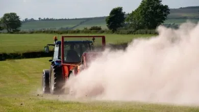 Tractor spreading fertilizer on a green field, creating a dust cloud.