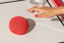Red Google Nest Mini speaker on a white table with a hand reaching towards it.