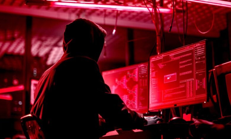 Person in hoodie types at computer screens in a dimly lit, red-toned room.