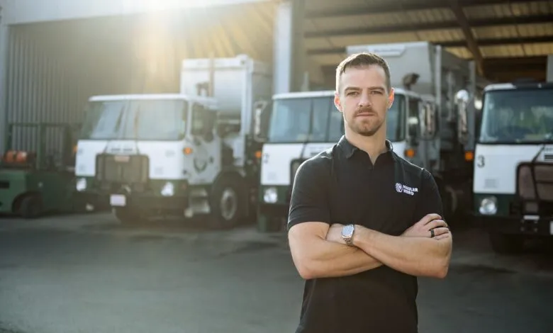 Man in black polo with arms crossed stands in front of garbage trucks.