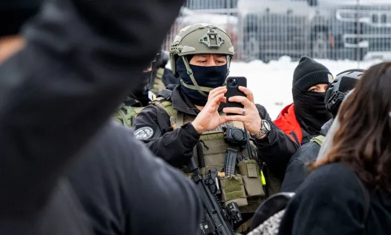 Officer in tactical gear takes photo with phone amidst crowd.