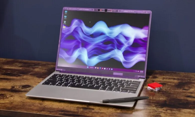 Open laptop on desk with stylus and red object, screen displays abstract purple design.