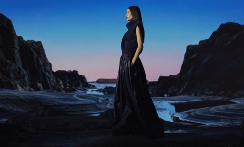Woman in dark outfit stands on rocky beach with blue and pink sky.