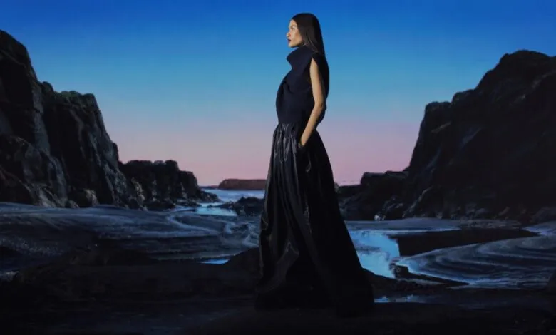 Woman in dark outfit stands on rocky beach with blue and pink sky.