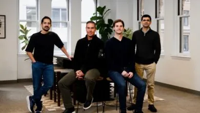 Four men pose indoors in a well-lit office space with large windows.