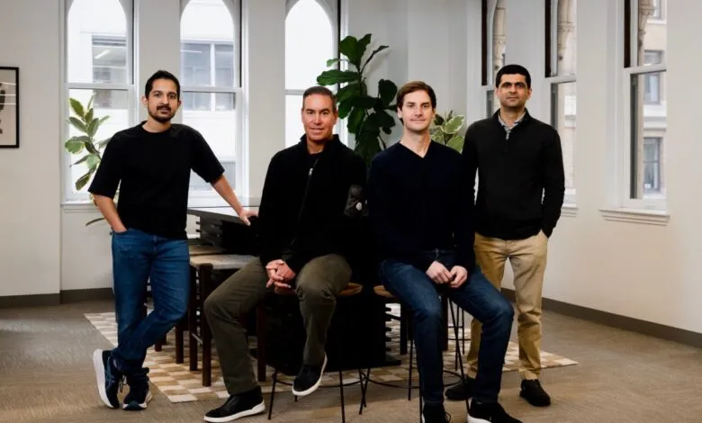 Four men pose indoors in a well-lit office space with large windows.