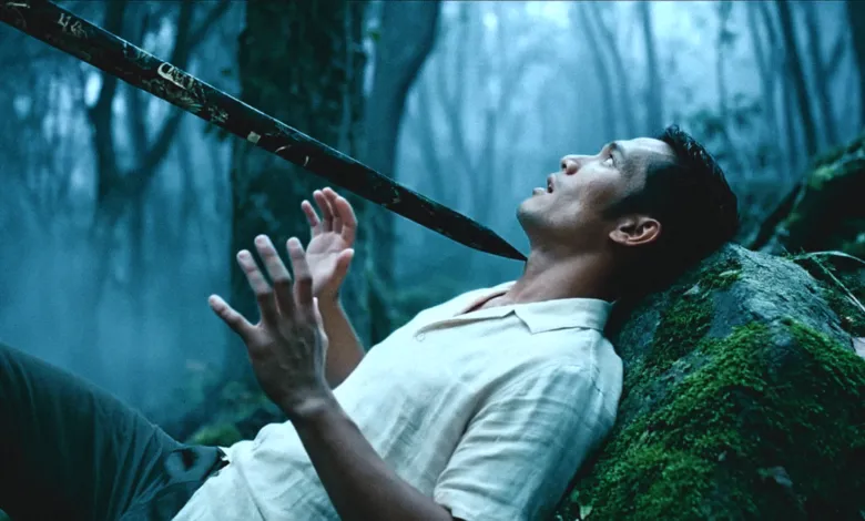 Man in forest with sword pointed at his neck, hands raised.